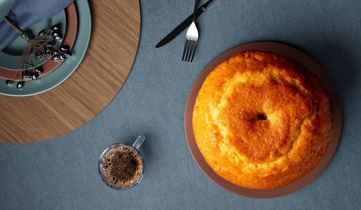 Fotografia de uma mesa elegantemente posta, com um bolo bundt dourado em um prato marrom. Ao lado, há pratos coloridos e um copo de café, com talheres dispostos de forma atraente.