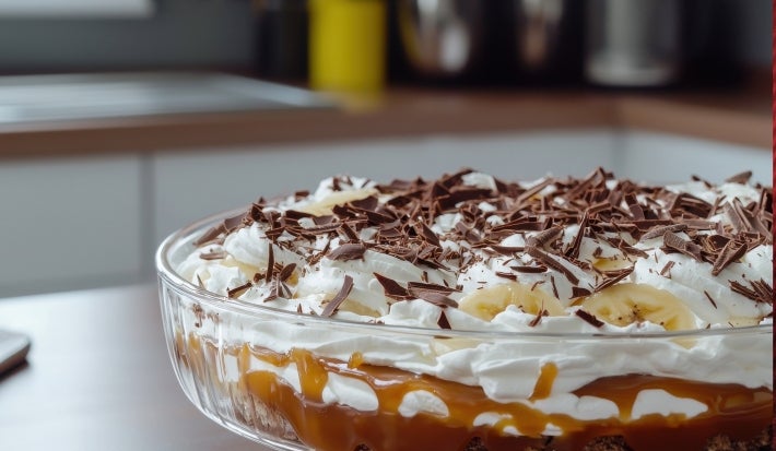 Em cima de uma mesa, refratário com biscoitos crocantes, doce de leite e chantilly, decorado com bananas em rodelas e raspas de chocolate.