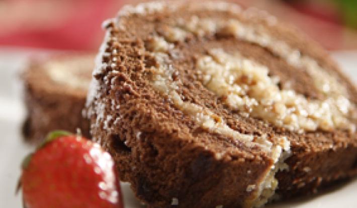Fotografia em tons de vermelho em uma bancada com uma toalha vermelha, um prato branco redondo raso e uma fatia de rocambole de chocolate recheado com coco e ao lado, um morango.
