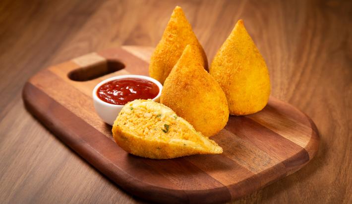 Fotografia em tom amadeirado com uma tábua de madeira ao centro. Em cima da tábua existe 3 coxinhas douradas e outra cortada pela metade recheada com frango desfiado. Ao lado existe um pequeno pote cheio de ketchup.