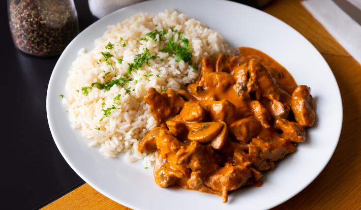 Foto aproximada da receita de strogonoff de carne tradicional, com um molho escuro marrom, pedaços de champignon, servido em um prato branco com arroz e ervas finas picadas