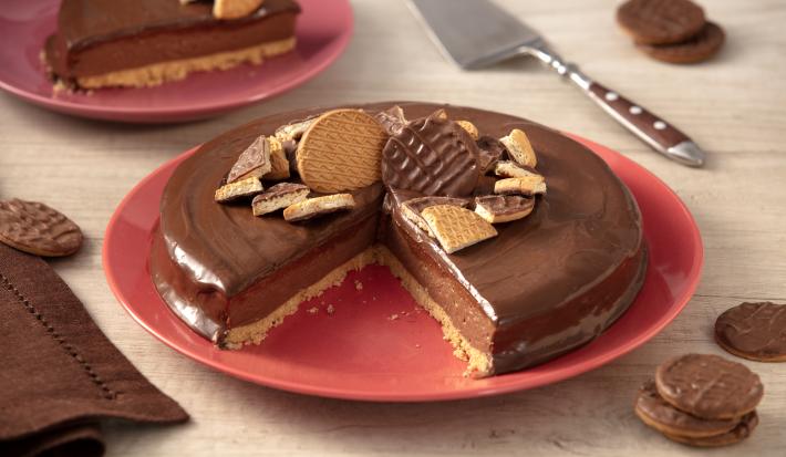 Foto de uma bancada clara. Sobre ela há um guardanapo de pano marrom, alguns Biscoitos Calipsos cobertos com chocolate espalhados e, ao centro, um prato vermelho com a receita pronta, sem uma fatia, mostrando o interior. Há um prato ao fundo com a fatia.