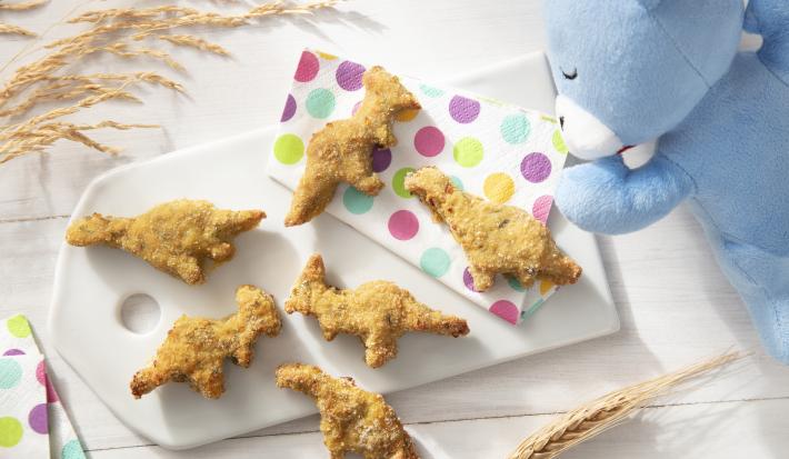 Foto da receita de Nuggets de Frango e Abobrinha. Observa-se uma tábua branca com os nuggets em formato de dinossauro sobre ela em cima de guardanapos coloridos de bolinha.