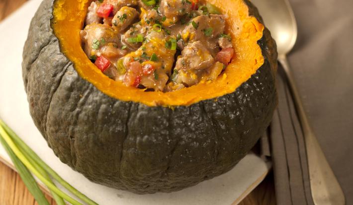 Fotografia em tons de marrom e cinza de uma bancada de madeira com um paninho branco, sobre ele uma abóbora recheada com carne e legumes. Ao lado cebolinhas e um paninho cinza com uma colher, ao fundo um prato branco.