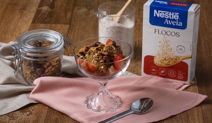 Fotografia de um recipiente decorativo de vidro com granola e alguns morangos cortados na metade, sobre um pano rosa de tom claro. Ao redor do recipiente tem uma embalagem de aveia flocos e um pote de vidro decorativo com a granola.