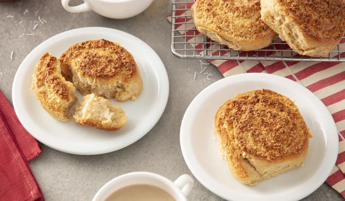 Foto da Receita de Coconut Rolls. Observa-se dois pratos de sobremesa com duas unidades de coconut rolls cada. Atrás, tem uma grelha com mais duas unidades. Duas xícaras de café com leite e toalhas vermelhas decoram a foto.