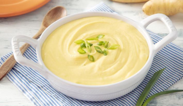 Foto em tons de amarelo da receita de purê de mandioquinha zero lactose servida em uma panela branca sobre um pano listrado azul e branco com uma colher de madeira ao lado
