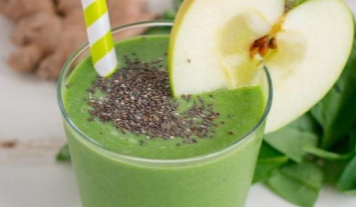 Fotografia em tons de verde em uma mesa branca com um copo de vidro alto e o smoothie de maçã verde com banana e chia dentro dele, com uma fatia de maçã enfeitando o copo. Ao fundo, folhas de espinafre e gengibre.