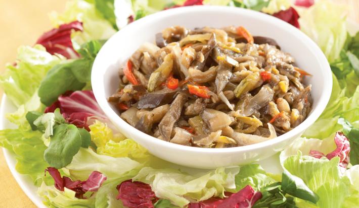 Fotografia em tons de verde e vermelho em uma bancada de madeira com um jogo americano amarelo. Ao centro, um prato branco raso redondo grande com salada de alface e abobrinha. E ao centro, um potinho com o antepasto de berinjela.