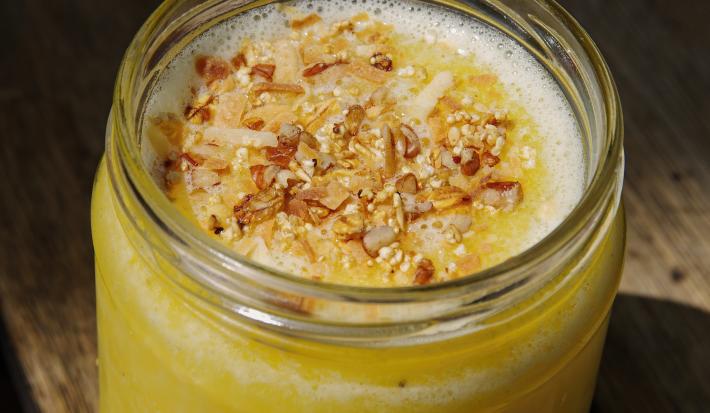Fotografia de um purê a base de maçãs e ameixas dentro de um recipiente decorativo de vidro, similar à um copo, com algumas nuts e pequenas lascas de coco por cima. O recipiente está sobre uma bancada de madeira.
