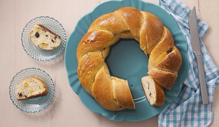 foto em tons de bege e azul de uma bancada bege vista de cima, contém um prato redondo azul com a rosca ao lado um pano xadrez azul e branco com uma faca. Do outro lado dois pratos azuis ambos com um pedaço da rosca com frutas secas.