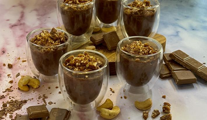 Foto da Receita de Mousse de Chocolate Crocante. Observa-se 6 taças de vidro individuais com a mousse dentro e com a pranilé de castanha de caju em cima decorando. Pedaços de chocolate e castanhas de caju decoram a foto também.