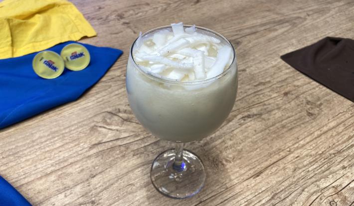 Fotografia em tons de amarelo e azul em uma bancada de madeira, com paninhos amarelo e azul e uma taça de vidro ao centro com a bebida de coco e Galak dentro, decorado com lascas de coco fresco.