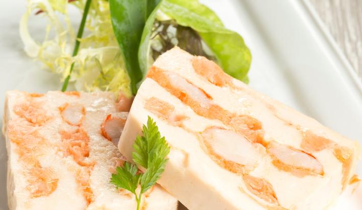 Fotografia de duas fatias de terrine de gorgonzola com nozes e cream cheese sobre um apoio branco retangular.