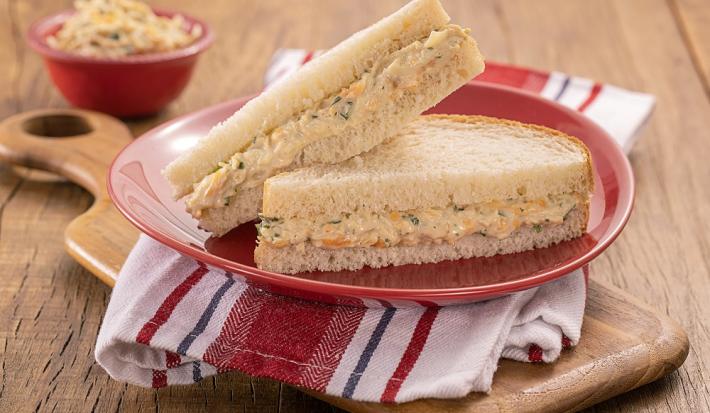 Foto da receita de sanduíche com patê de frango servido em duas porções juntas em um prato vermelho sobre um pano branco e vermelho listrado. Embaixo uma tábua de madeira e ao fundo um recipiente vermelho com o frango desfiado para decoração