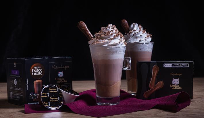 Fotografia em tons escuros de duas xícaras de vidro longas com Chococcino Mousse Língua de Gato, chantilly e língua de gato sobre um pano vinho. Ao lado tem uma caixa de nescafé e a outra de língua de gato. .