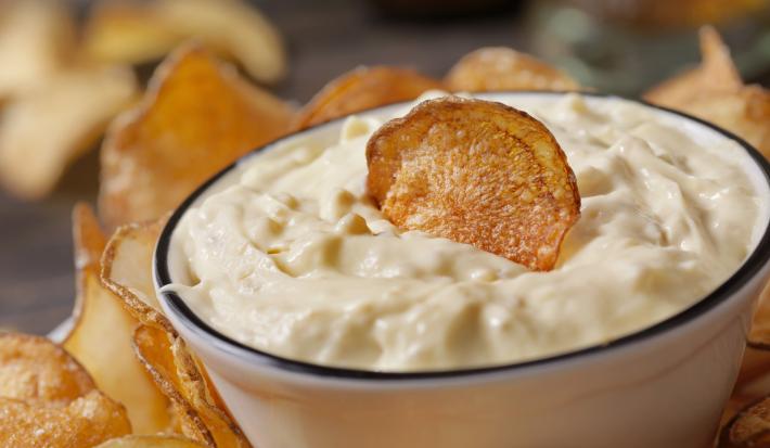 Fotografia de um recipiente pequeno de vidro com um patê de creme de leite, ketchup e creme de cebola com um chip de batata por cima. Em volta e no fundo, mais chips de batata sobre um apoio preto.