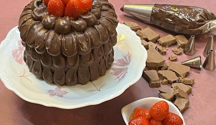 Foto em tons de marrom e vermelho da receita de bolo confeitado com ganache com morangos em cima, servido em um prato grande de porcelana branco com adornos em rosa. Ao lado, pedaços de chocolate e um bowl branco com morangos, além de um saco de confeitar