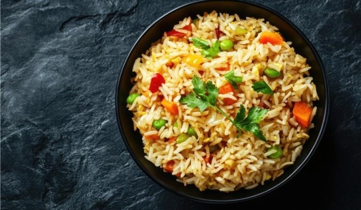 Em um prato há arroz soltinho com uma mistura de legumes, como cenoura, ervilhas e pimentões, decorado com folhas de salsinha.