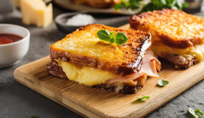 Fotografia de dois lanches de queijo e presunto com mais queijo gratinado por cima de cada um, que estão juntos sobre uma tábua de madeira.