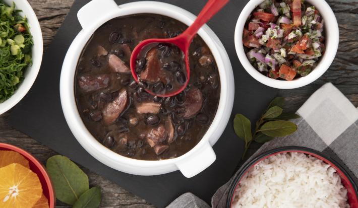 Imagem vista de cima de uma travessa redonda com feijoada, ao lado um pote de vinagrete, um pote de arroz, um pote de laranja e um pote de couve.
