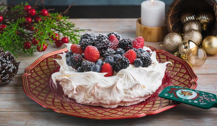 Foto da receita de pavlova natalina, branca, sobre um prato vermelho, decorada com frutas vermelhas e açúcar. Ela está numa mesa de madeira preenchida de decorações de natal como pinhas, bolas de natal e uma vela acesa ao fundo