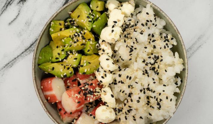 Fotografia em tons claros com um bowl ao centro. Dentro do bowl existe uma refeição a base de arroz e kani, temperada com gergelim e molho