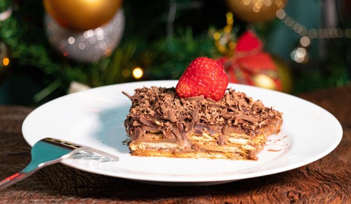 Fotografia de um pedaço de pavê de damasco com chocolate, cobertura de ganache, raspas de chocolate e um morango por cima. O pavê está em um prato raso branco, o qual está sobre uma mesa de madeira.