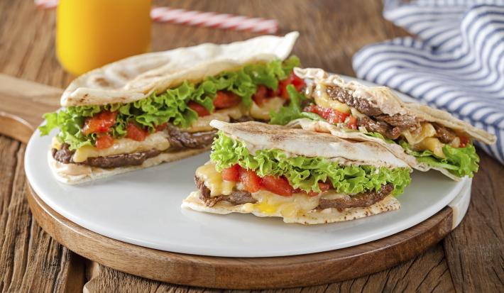 Fotografia em tons de branco e verde de bancada de madeira, tábua redonda com beirute partido ao meio, e uma das metades partidas ao meio mostrando a carne, queijo, tomate, alface e pão sírio. Ao fundo copo com suco laranja e pano listrado azul e branco.