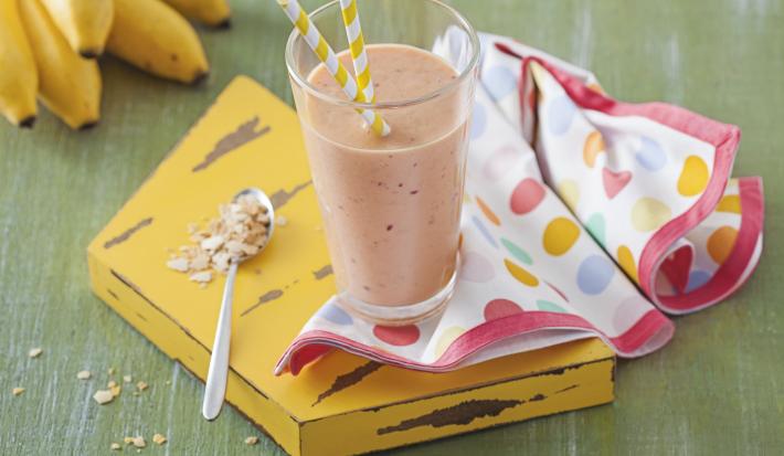 Fotografia em tons de verde e amarelo de uma bancada verde com uma tábua amarela, sobre ela um paninho com bolinhas coloridas e um copo com vitamina de frutas. Ao lado uma colher. Ao fundo um cacho de banana.