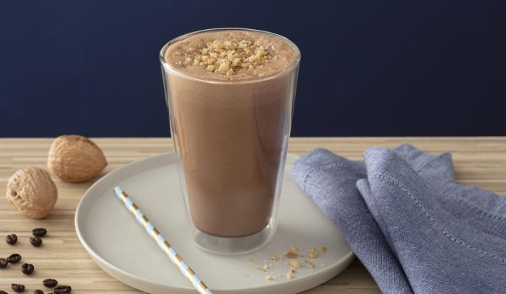 Foto em tons de marrom da receita de smoothie de café com nozes servida em um copo alto sobre uma base de porcelana cinza com um canudo listrado ao lado. Como decoração há duas nozes e um paninho azul