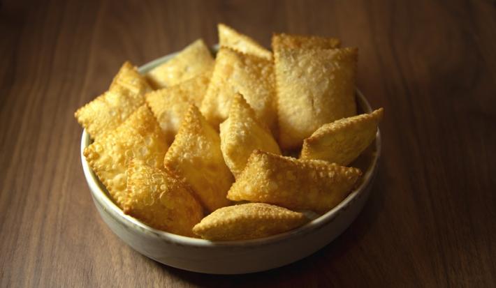 Fotografia em tons de amarelo, em uma bancada de madeira de cor marrom. Ao centro, um recipiente branco contendo os pasteis.