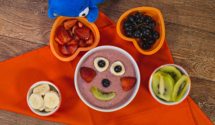 Fotografia em tons de laranja em uma bancada de madeira escura, um pano laranja, um recipiente redondo com o smoothie de morango, banana e kiwi dentro dele e frutinhas formando um rostinho feliz. Ao lado, potinhos com as frutas e o ursinho azul de pelúcia