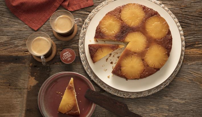 Foto de cima de um bolo invertido com rodelas de abacaxi, uma fatia está servida em um prato de sobremesa ao lado e duas xícaras transparentes com café preto e uma cápsula de Dolce Gusto Café Matinal.