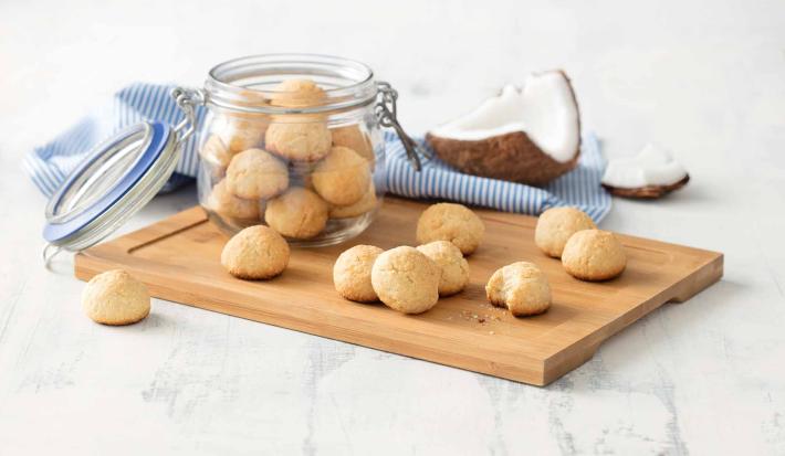 Receita em tons de bege e azul de uma bancada branca vista de cima. Contém uma tábua bege com um pote transparente com biscoitinhos dentro e por cima da tábua contém mais biscoitos. Ao fundo um pano azul e uma coco cortado.