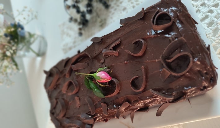 Fotografia do bolo de banana, chocolate e aveia assado em uma forma de bolo inglês, sobre um apoio retangular branco. O bolo tem uma cobertura de brigadeiro e decorado com raspas de chocolate.