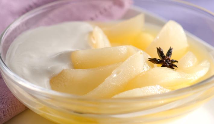 Fotografia em tons de roxo em uma bancada de madeira com toalha roxa, um paninho lilás, um recipiente redondo de vidro com as peras com creme de leite dentro dele.