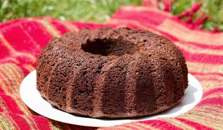 Fotografia de um bolo de chocolate em forma de bundt, com uma cor marrom profunda e textura densa, colocado em um prato branco. O fundo é um tecido listrado em vermelho e verde.