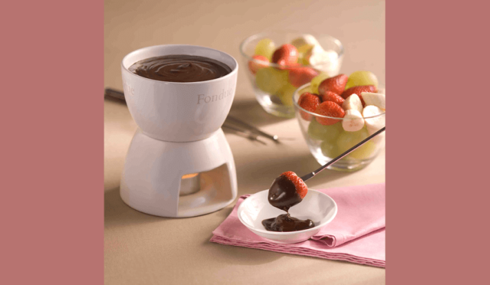 Fotografia de uma panela de fondue de cerâmica branca e aquecida com uma vela abaixo. Ao lado frutas frescas como morango, uva e banana em dois recipientes de vidro e um garfo próprio para fondue com uma morango coberto de chocolate.