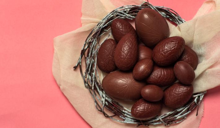 fotografia em tons de rosa e marrom de uma bancada rosa vista de cima, contém um cesto redondo com mini ovinhos de chocolate