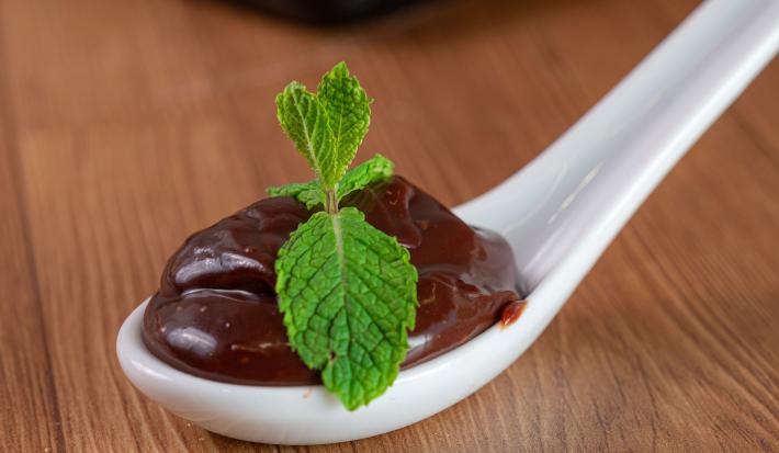 Fotografia de uma colher de servir branca de vidro, com uma porção de brigadeiro com duas folhas de hortelã por cima. A colher está apoiada sobre uma mesa de madeira.