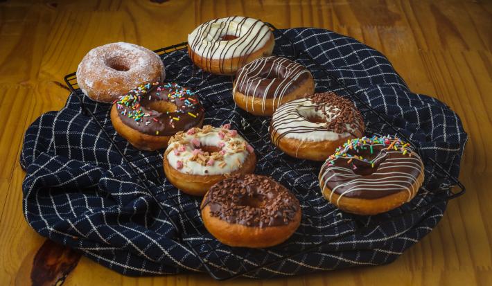 Foto aproximada de uma bancada de madeira com uma grade coberta por um tecido azul quadriculado e sobre ele há oito donuts, alguns decorados com chocolate branco, outros com chocolate ao leite e alguns com confeitos comestíveis