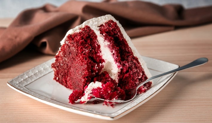 Fotografia de uma fatia de bolo veludo vermelho em um prato cerâmico branco, com creme de queijo, e um garfo prateado ao lado. O fundo é um tecido marrom suave, sobre uma mesa de madeira clara.