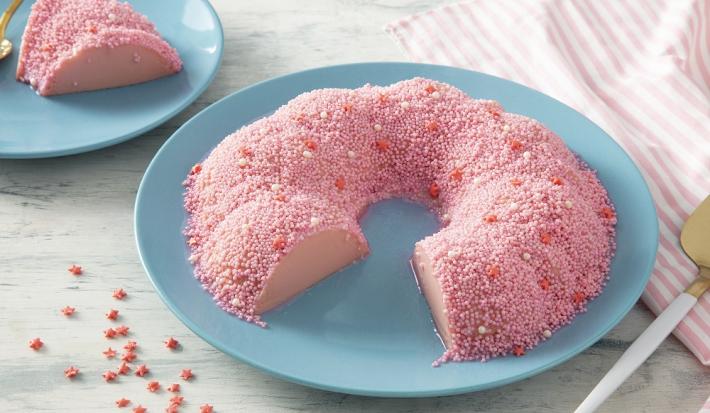 Foto em tons de cor de rosa da receita de pudim rosa servida em uma porção grande com uma fatia cortada sobre pratos de porcelana azul claro