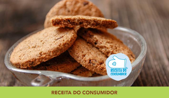 Foto aproximada da receita de Cookies Integrais, dentro de um pote de vidro, em tom marrom claro, sobre uma bancada de madeira e com dizeres no rodapé em tom verde.
