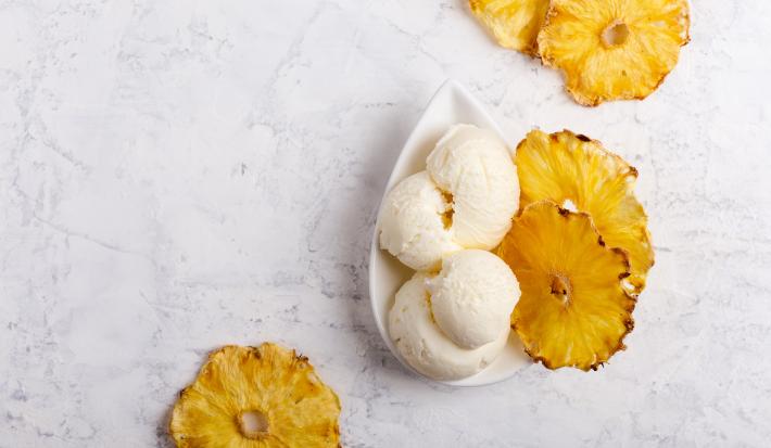 fotografia em tons de cinza, branco e amarelo de uma bancada cinza vista de cima. Contém um recipiente oval branco com bolas de sorvete e em volta fatias de abacaxi.