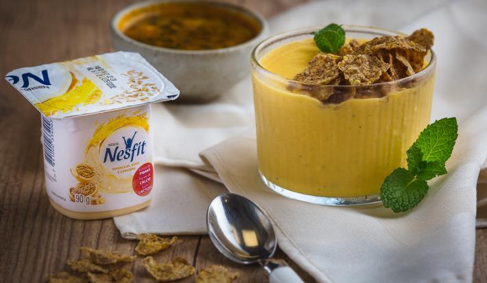 Fotografia em tons de amarelo em uma bancada de madeira, um paninho bege, um potinho com um creme de iogurte de maracujá com gengibre. Ao lado, um potinho de maracujá e uma colher.