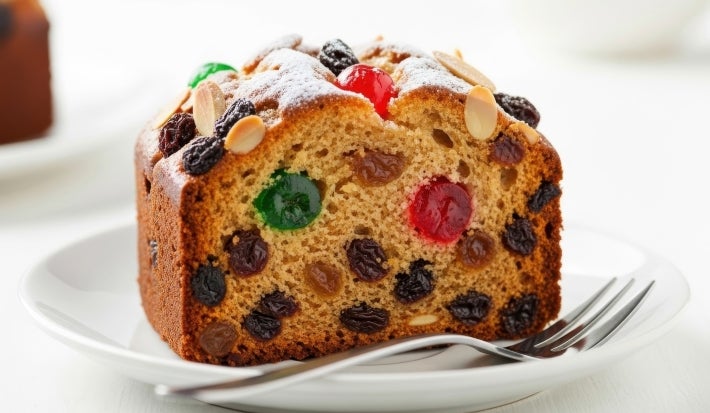 Fotografia de uma fatia de bolo de frutas em um prato branco, com uma crosta dourado-marrom e recheada com frutas cristalizadas, passas, decorado com amêndoas e polvilhada com açúcar de confeiteiro.