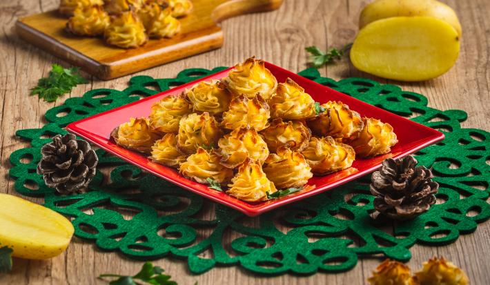 Foto da receita Batatas Natalinas, em tom amarelo e dourado, servidas em um prato vermelho quadrado sobre uma toalha verde natalina. Tudo está em uma mesa de madeira decorada com batatas cruas, pinhas natalinas e mais algumas batatas prontas.