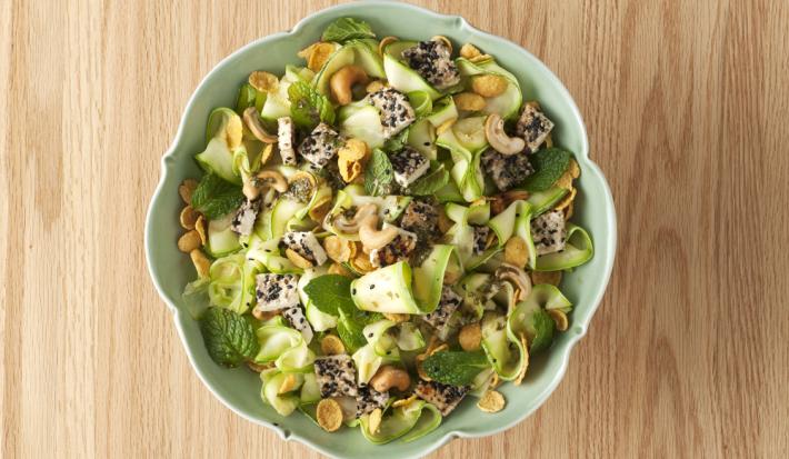 fotografia em tons de bege e verde de uma bancada vista de cima. Ao centro contém um recipiente redondo com salada de abobrinha.